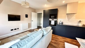 A modern living room with a sofa, a flat-screen TV, a kitchen area with appliances, and a dining table. - Redland Suites - Apartment 7 (Redland)