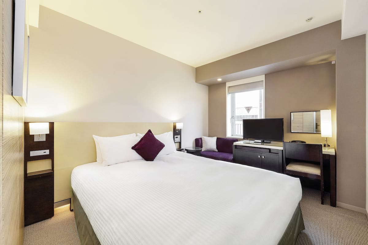 Ein Hotelzimmer mit einem großen Bett, einem Schreibtisch mit Stuhl, einem Fernseher und einem Fenster mit Jalousien.