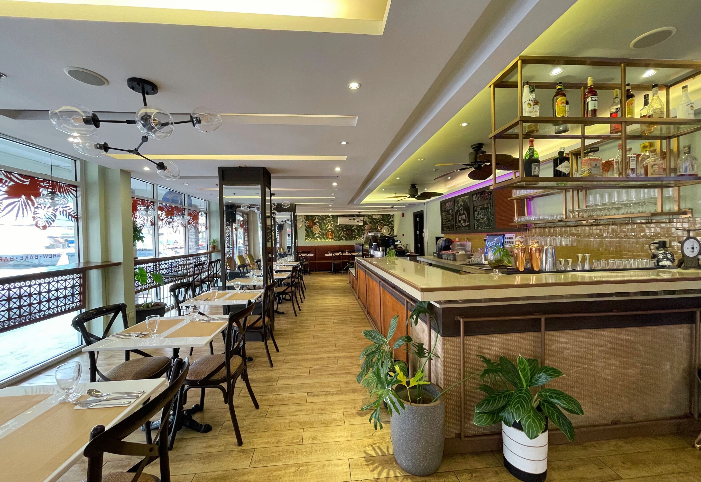 A restaurant interior with wooden flooring, tables with chairs, a bar area with shelves of bottles, and a large window with decorative glass panels.