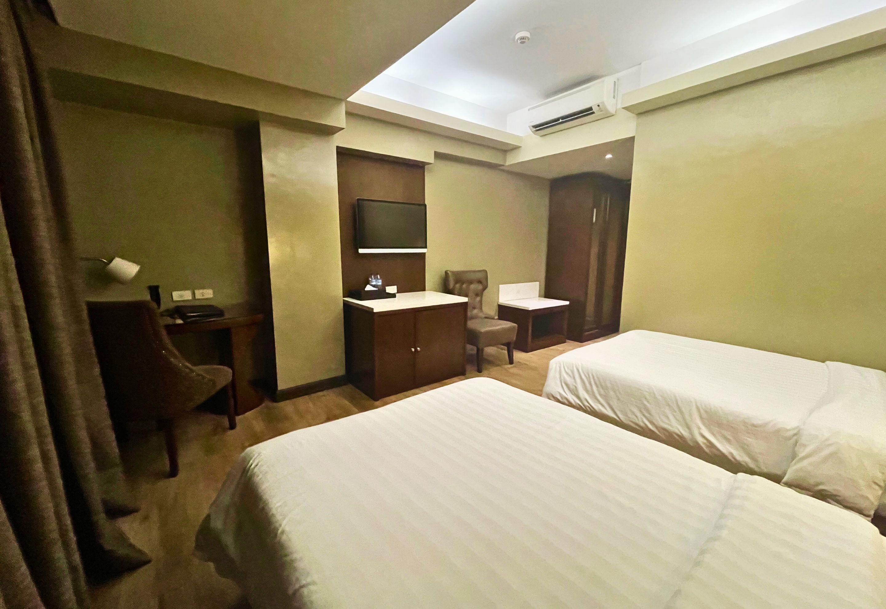 A hotel room with two beds, a desk, a chair, a TV, and an air conditioning unit.