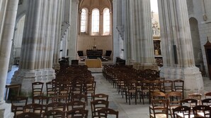 Interior de uma catedral com altos tetos abobadados, grandes colunas de pedra e fileiras de bancos de madeira.