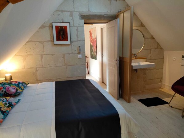 A bedroom with a bed, a bedside table with a lamp, a wooden door leading to another room, and a small wooden shelf with a mirror. - Les Logis d Ussé (Rigny-Usse)