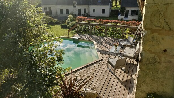 Um deck de madeira com piscina, rodeado por vegetação e um edifício branco ao fundo.