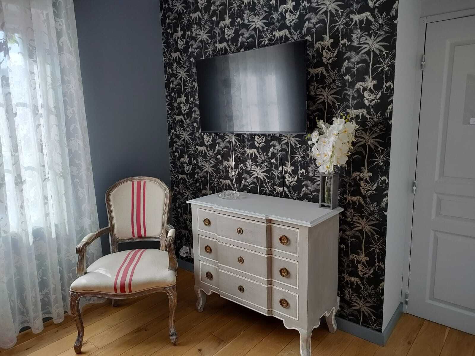 A room with a wooden floor, a white chair with red stripes, a white dresser with gold knobs, and a TV mounted on the wall.