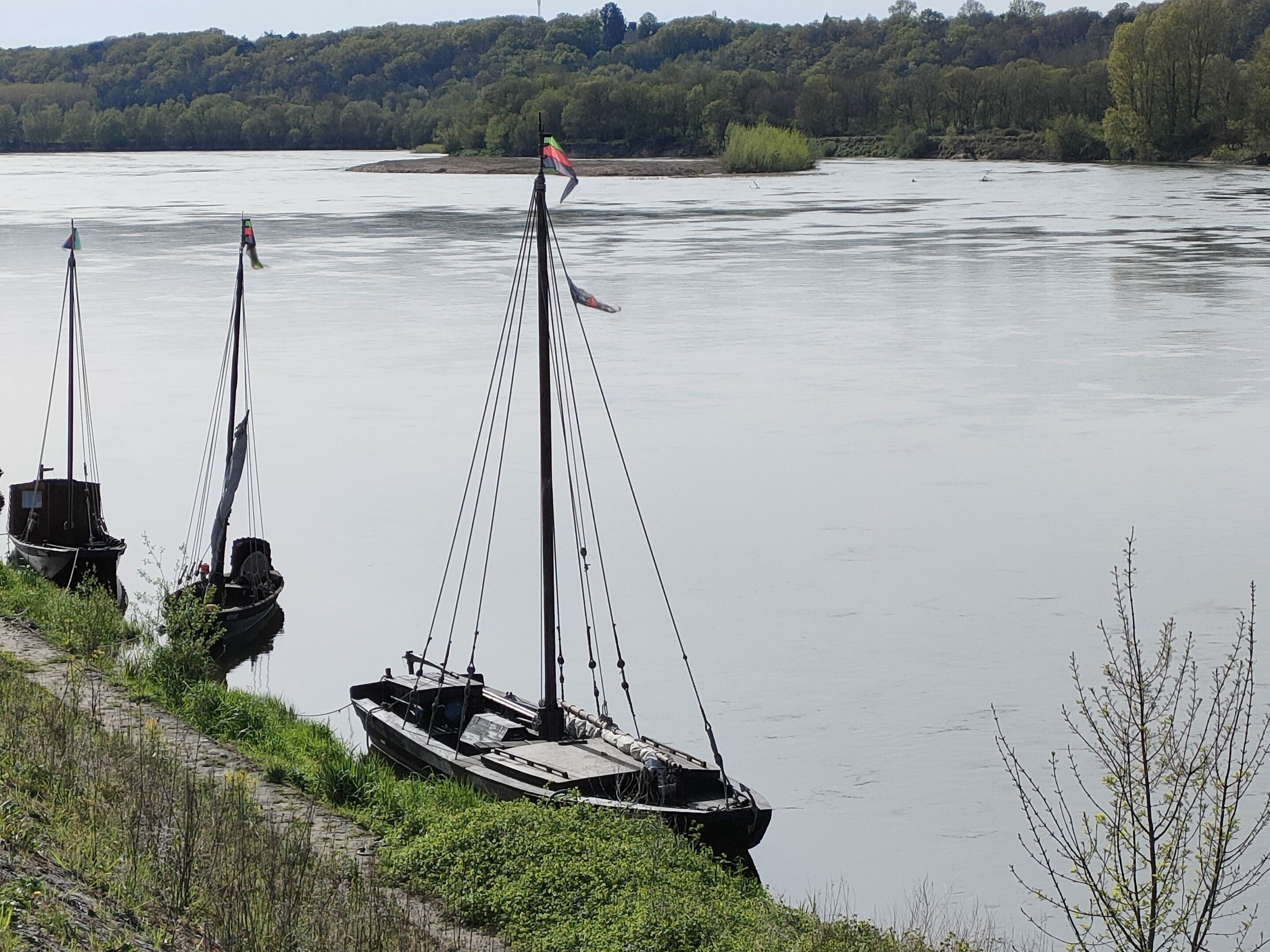 Trois bateaux sont amarrés le long d’une rive, avec une colline boisée en arrière-plan.