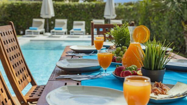 Une table à manger avec des assiettes, des verres de jus d’orange et un plateau de fruits, le tout placé près d’une piscine.