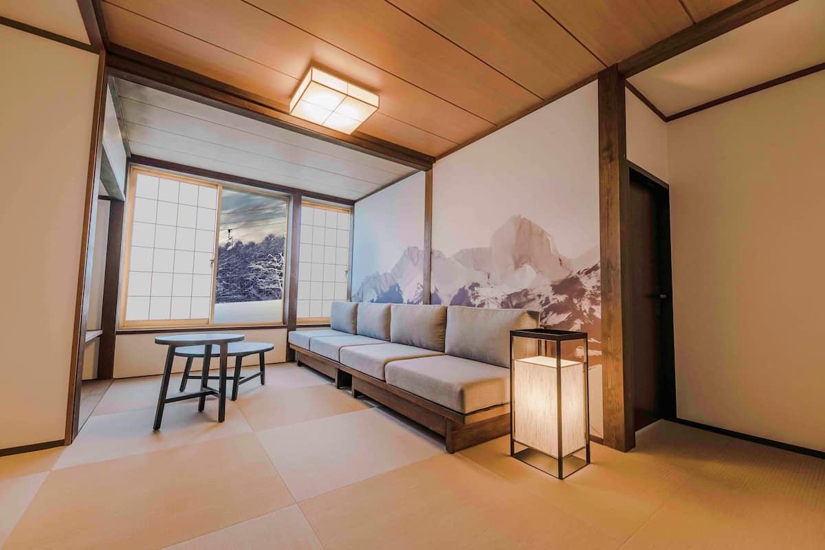 A room with tatami flooring, a low wooden ceiling, a sofa, a table, and a wall-mounted projector displaying a mountain scene.