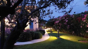 Un vialetto ben illuminato conduce a un edificio con una porta blu, circondato da verde lussureggiante e piante fiorite.