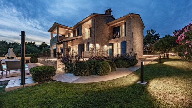 Una moderna casa a due piani con facciata in pietra, un balcone e un'area esterna ben illuminata con piscina e posti a sedere.