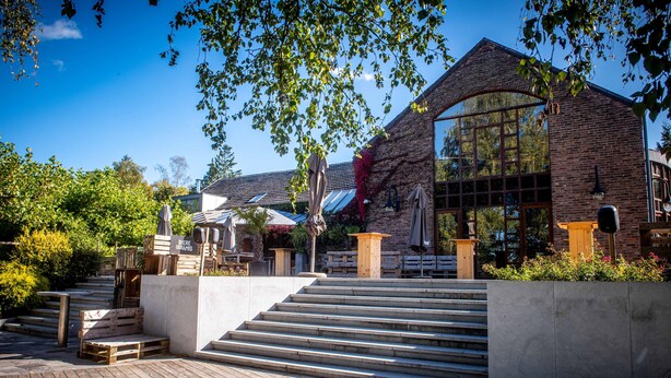 A building with a brick facade, large windows, and a wooden deck leading to the entrance. - Aux Etangs - Hôtel (Herve)