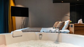 A modern hotel room with a large bed, a bathtub with bubbles, a flat-screen TV, and a wooden side table. - Aux Etangs - Hôtel (Herve)