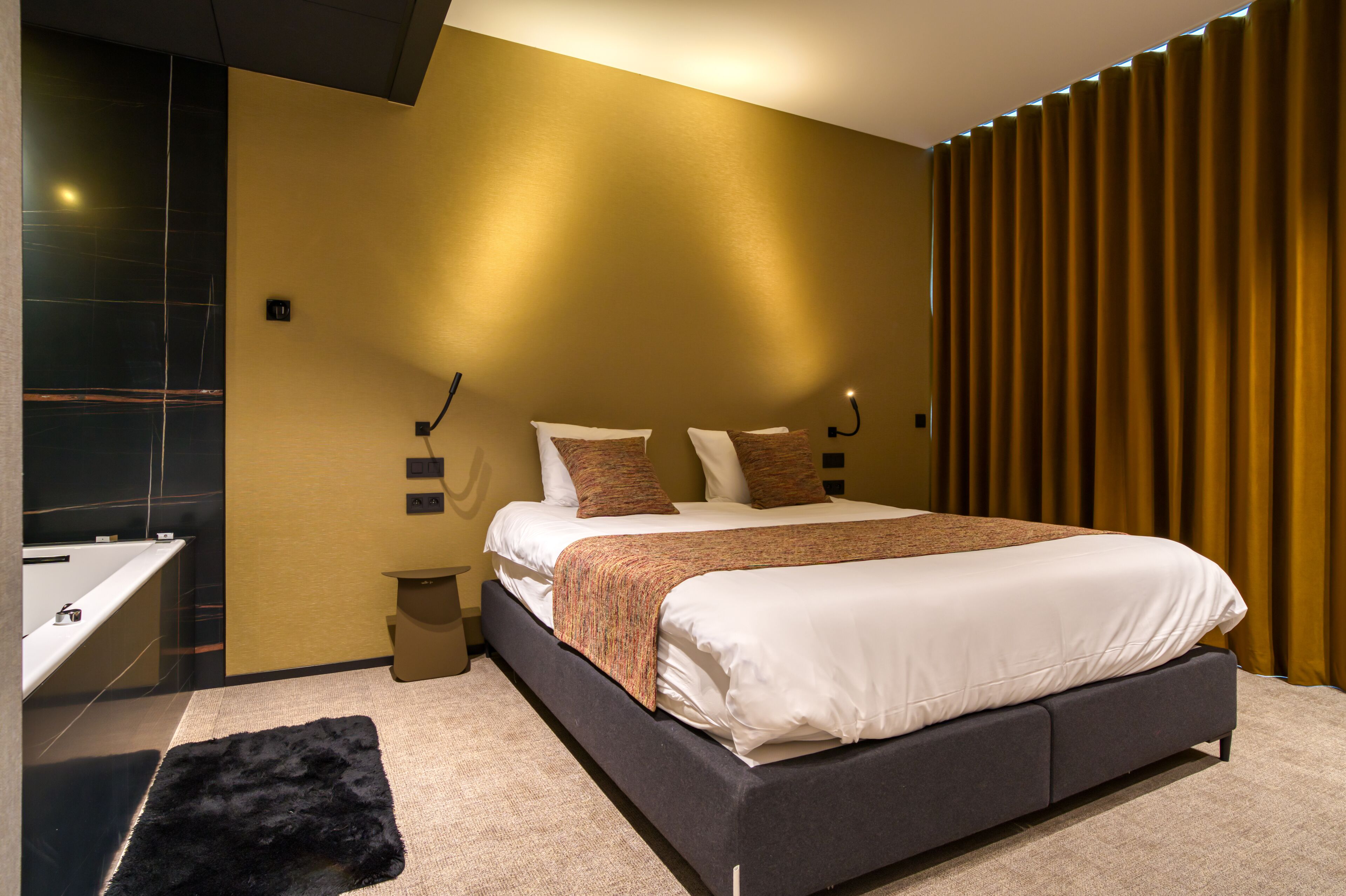 Una habitación de hotel moderna con una cama grande, una mesita de noche, una lámpara de noche pequeña, un tapete negro y un baño con un lavabo y espejo blancos.