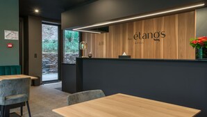 A modern hotel reception area with a wooden reception desk, a seating area with cushioned chairs, and a view of greenery outside. - Aux Etangs - Hôtel (Herve)