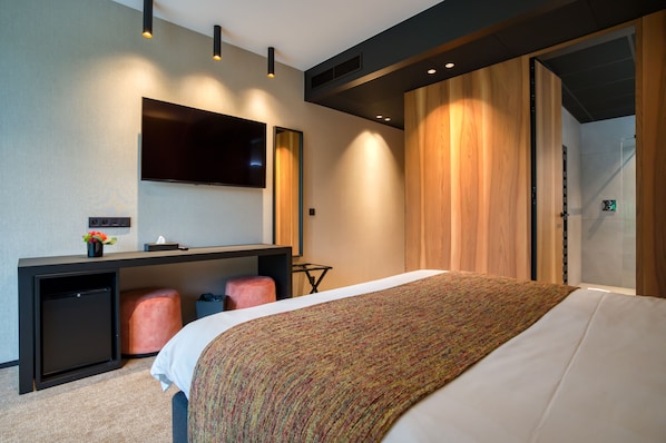 A modern hotel room with a large bed, a desk with a flat-screen TV, a small fridge, and a wooden wardrobe.