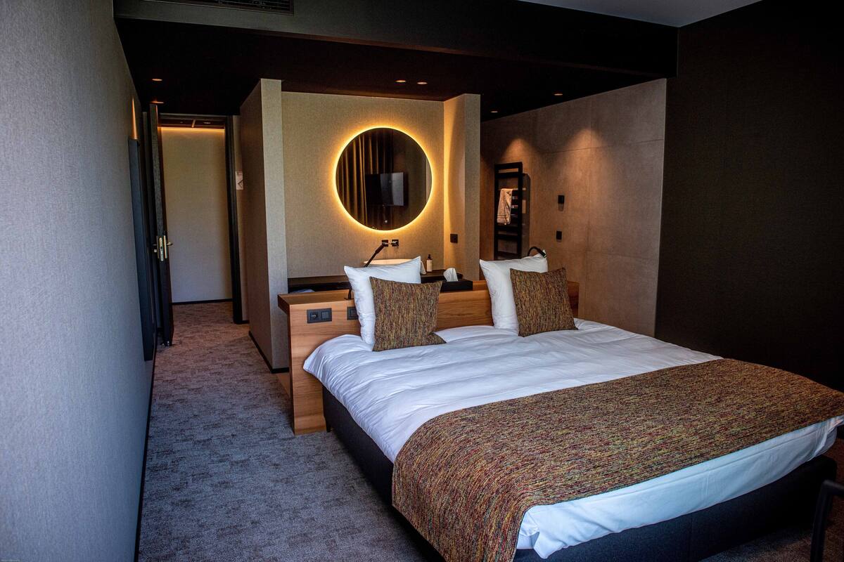 A modern hotel room with a large bed, a round mirror, a wooden nightstand, and a small table with a lamp.