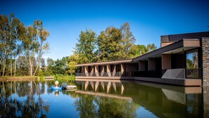 A modern building with a reflective water body in front, surrounded by trees. - Aux Etangs - Hôtel (Herve)