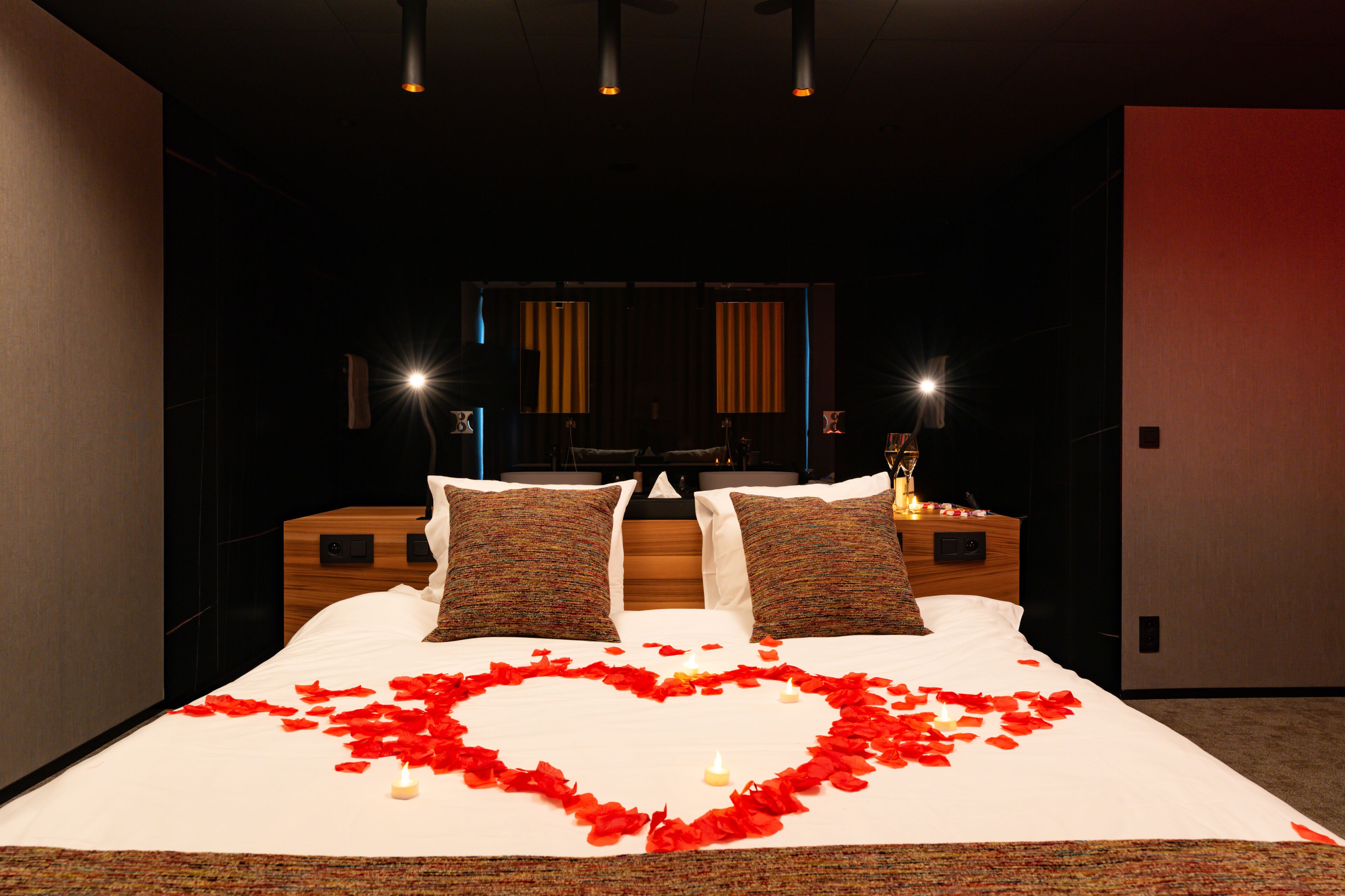 Habitación de hotel con una cama grande, dos mesitas de noche, un espejo y una decoración en forma de corazón rojo sobre la cama.