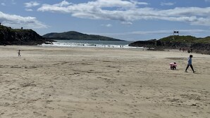 Ein weiter Sandstrand mit Menschen, die spazieren gehen und spielen, Hügel im Hintergrund und einem klaren Himmel.