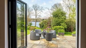 Eine Terrasse mit Korbmöbeln und Steinboden, mit Blick auf einen See und Grünflächen.