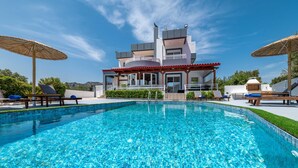 A modern pool area with a clear blue pool, lounge chairs, umbrellas, and a contemporary building with balconies. - Villa Di Georgia (Αφάντου)