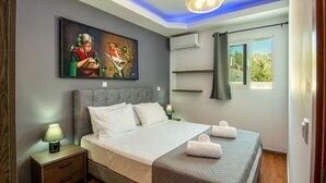 A bedroom with a bed, nightstands, a painting, an air conditioner, and a window with curtains. - Villa Di Georgia (Αφάντου)