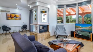 A modern living room with a fireplace, comfortable seating, and large windows offering scenic views. - Villa Di Georgia (Αφάντου)