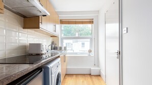 A modern kitchen with a built-in oven, a washing machine, and a window with a view of a house. -  Cosy studio Apartment near Hyde Park (London)