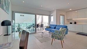 A modern living room with a blue sofa, a coffee table, and a patterned armchair. A kitchen with white cabinets is visible in the background. - Beautiful View! 2 bdr/ 2 bthr, beach100m and 300m Old Town, pool, garage, aircon (Albufeira Old town)