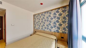 Una habitación pequeña con una cama individual, cabecera de madera, ventana con cortinas y una puerta.