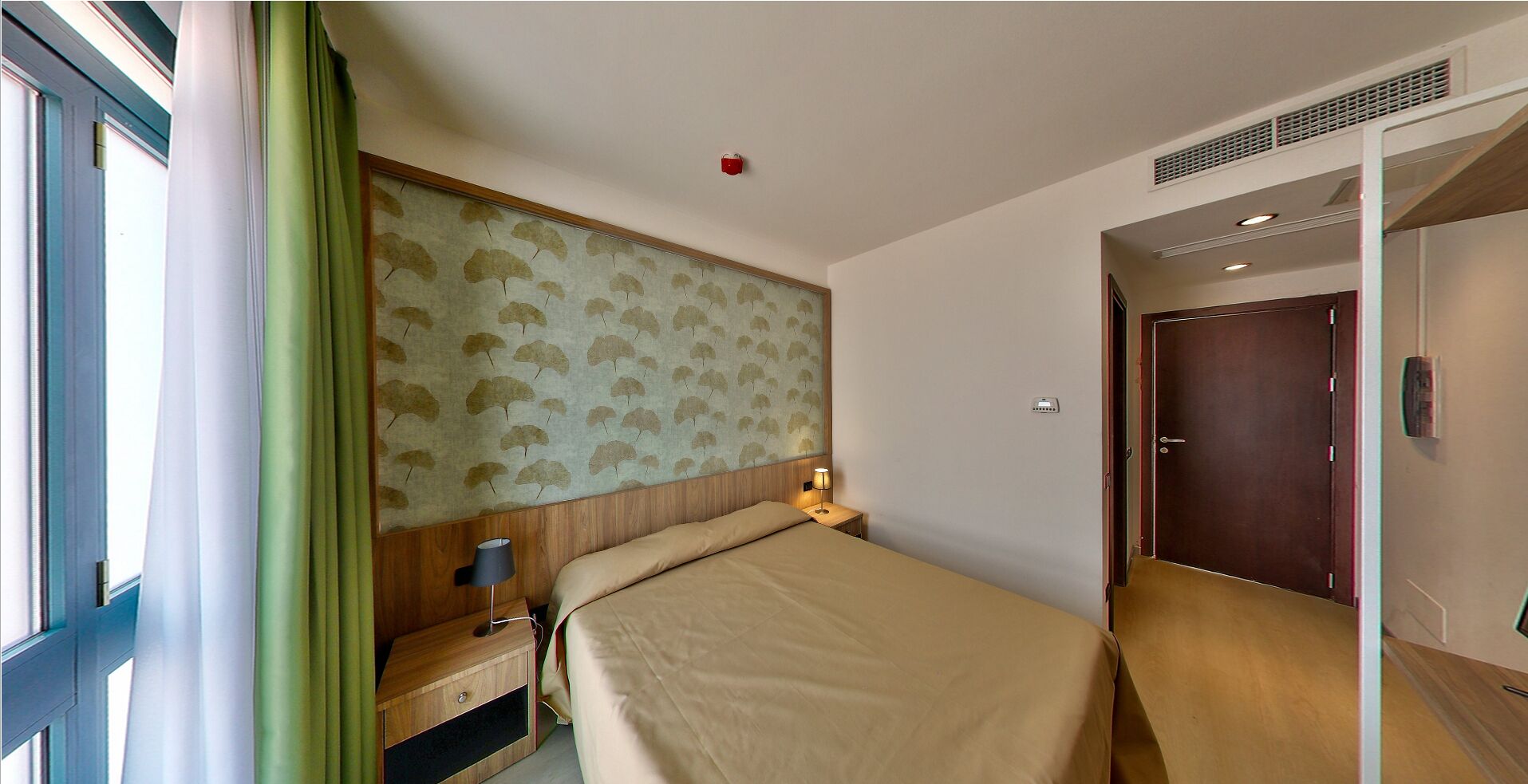A hotel room with a bed, a nightstand, a lamp, a wall with a leaf pattern, a door, and a window with curtains.
