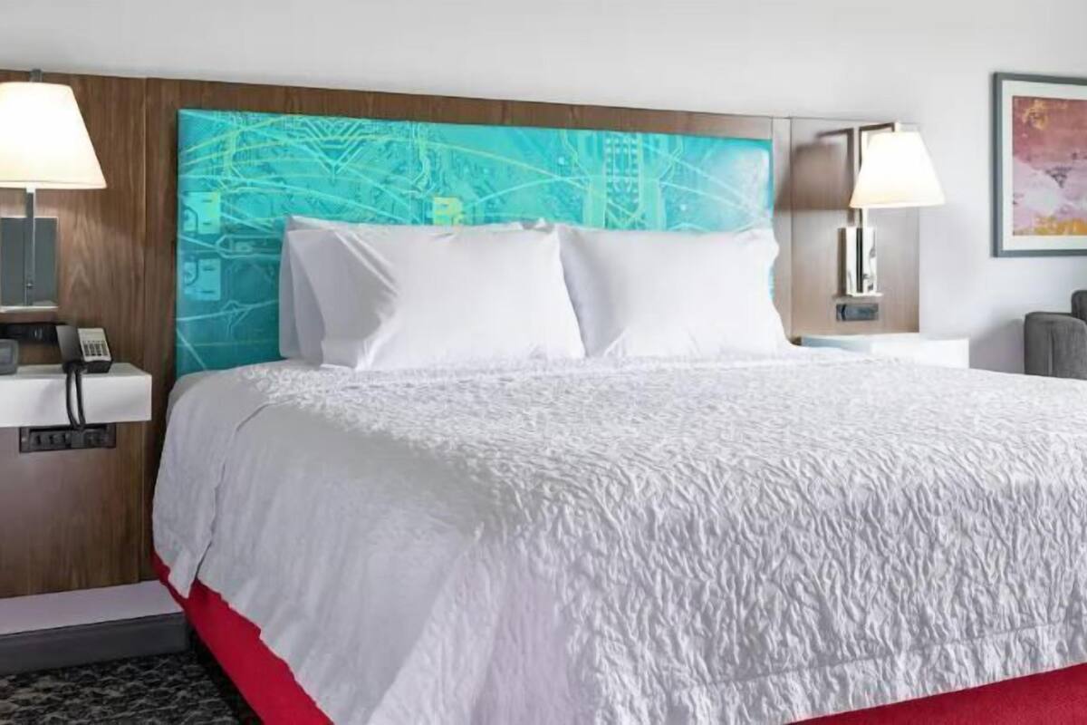 A hotel room with a large bed, two bedside tables with lamps, a chair, and a wall with a cityscape painting.