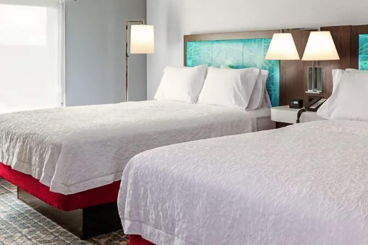 Two double beds with white linens, a wooden headboard with a blue abstract design, and bedside lamps.