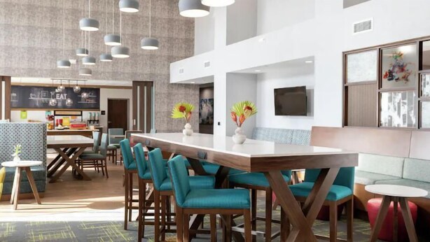 A modern restaurant with a long communal table, teal chairs, and a bar area with stools. - Hampton Inn Tappahannock (Tappahannock)