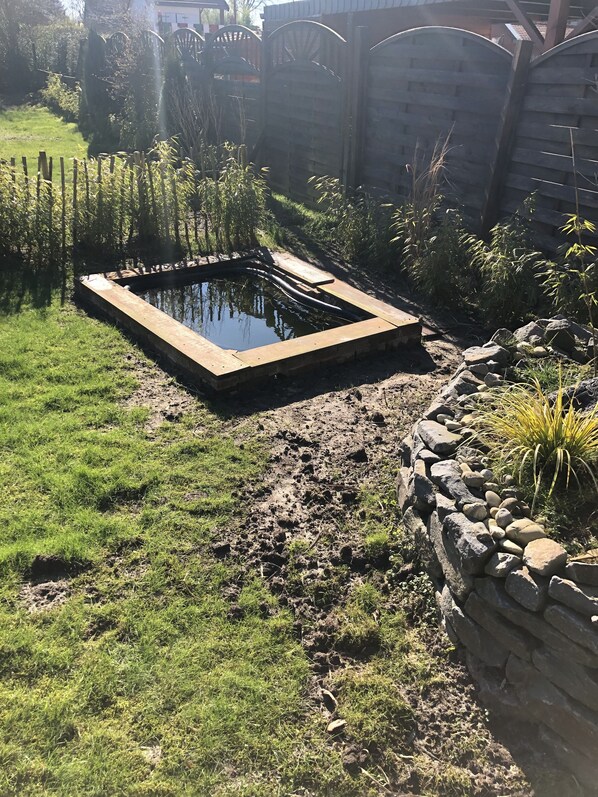 Un jardín con un estanque rectangular, un muro de piedra y una valla de madera.