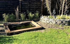 Ein rechteckiger Teich mit Steinrand, umgeben von Gras und Bambuspflanzen, mit Holzzaun und einer Steinmauer im Hintergrund.