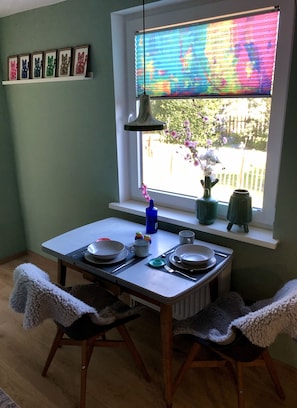 Ein Essbereich mit Tisch für zwei Personen, einem Fenster mit bunter Jalousie und einer Vase mit Blumen auf dem Fensterbrett.
