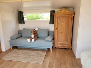 Ein Zimmer mit grauem Sofa, einem Holzkasten und einem Fenster mit Vorhängen.