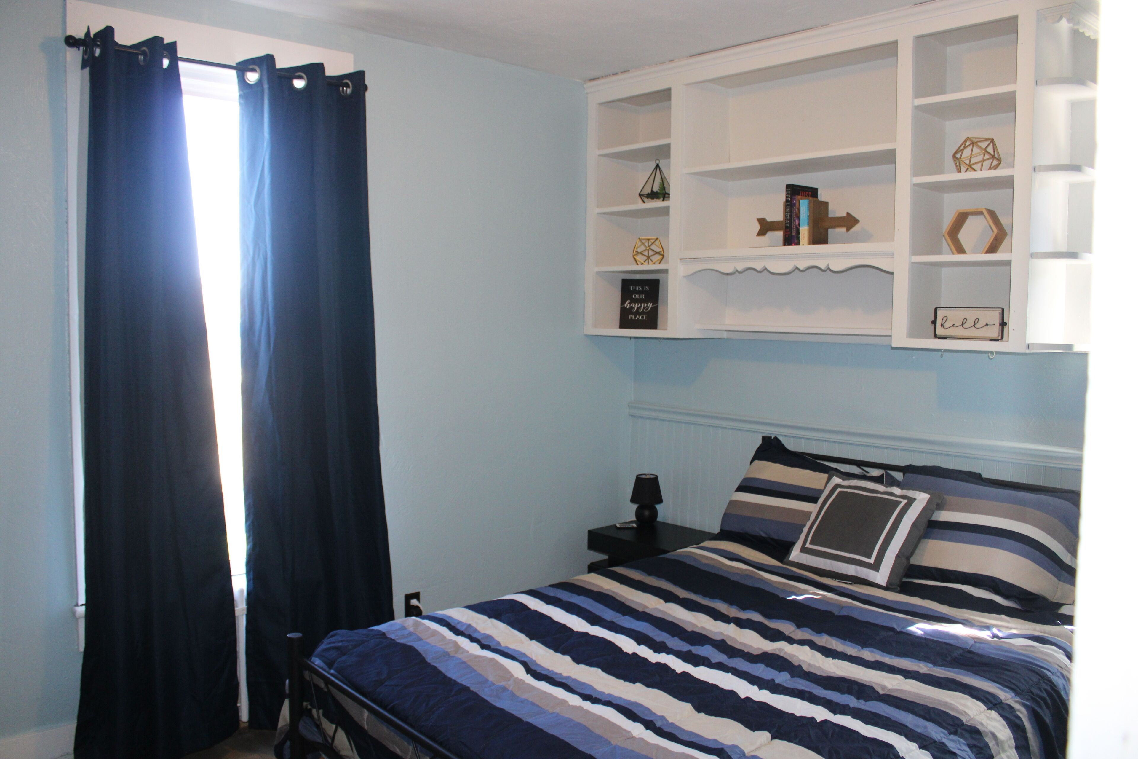 A bedroom with a bed, a nightstand, a shelf with decorative items, and a window with curtains.