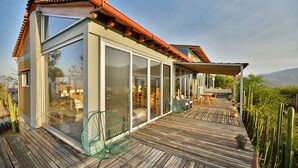 Una casa moderna con grandes ventanales, una terraza de madera y un patio con sillas y una mesa.