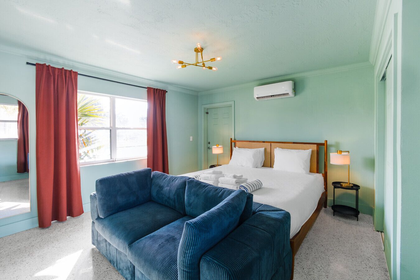 Une chambre d’hôtel avec un lit, un canapé bleu, un lustre et une fenêtre avec des rideaux.