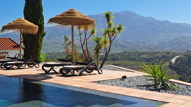 Zona de piscina con sillones de descanso y sombrillas de paja, rodeada de un paisaje montañoso.