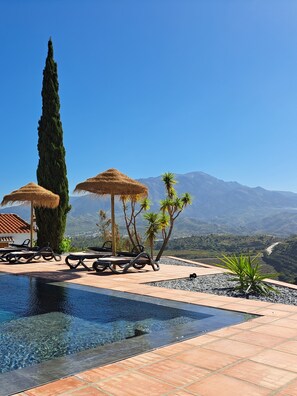 A pool area with lounge chairs and thatched umbrellas, surrounded by a mountainous landscape. - Villa with a private pool, sleeps 10, 5 bathrooms (Viñuela)