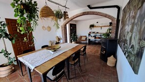 Un comedor rústico con una mesa de madera, sillas, plantas colgantes y un arco de ladrillo que da a una cocina.