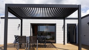 A covered outdoor seating area with a table and chairs, a wooden deck, and a glass door leading to an interior space. - Chalet for 6 in an estate with swimming pool (Bretignolles-sur-Mer)