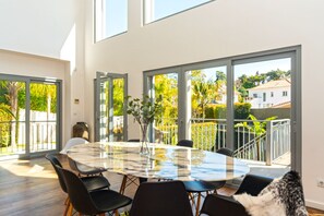 Un espace de repas moderne avec une table et des chaises en verre, de grandes fenêtres et une vue sur la verdure et les bâtiments environnants.