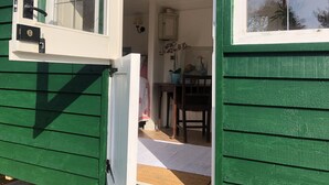 Une maison en bois peinte en vert, avec une porte et une fenêtre blanches. À l’intérieur, on trouve une cuisine avec une table, des chaises et une plante en pot.