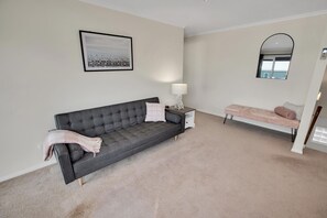 Una habitación con un sofá gris, una mesita blanca con una lámpara, un banco rosa y un cuadro en la pared.
