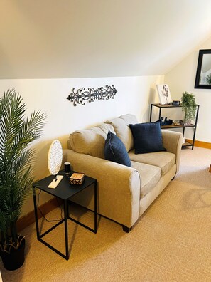 A living room with a beige sofa, a small black side table, a decorative wall piece, and a potted plant.