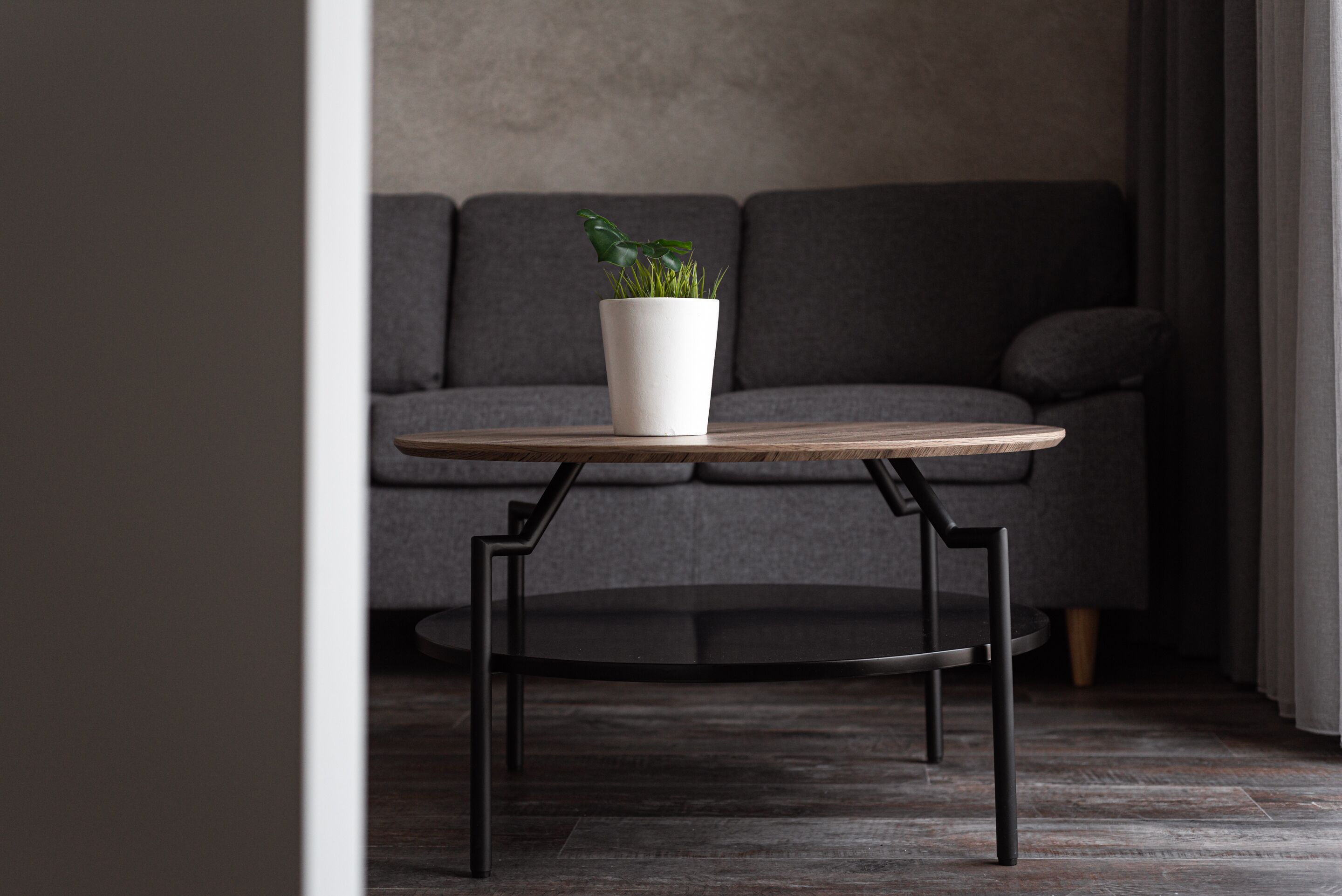 Ein runder Holztisch mit einem weißen Pflanzgefäß darauf, vor einer grauen Couch platziert.