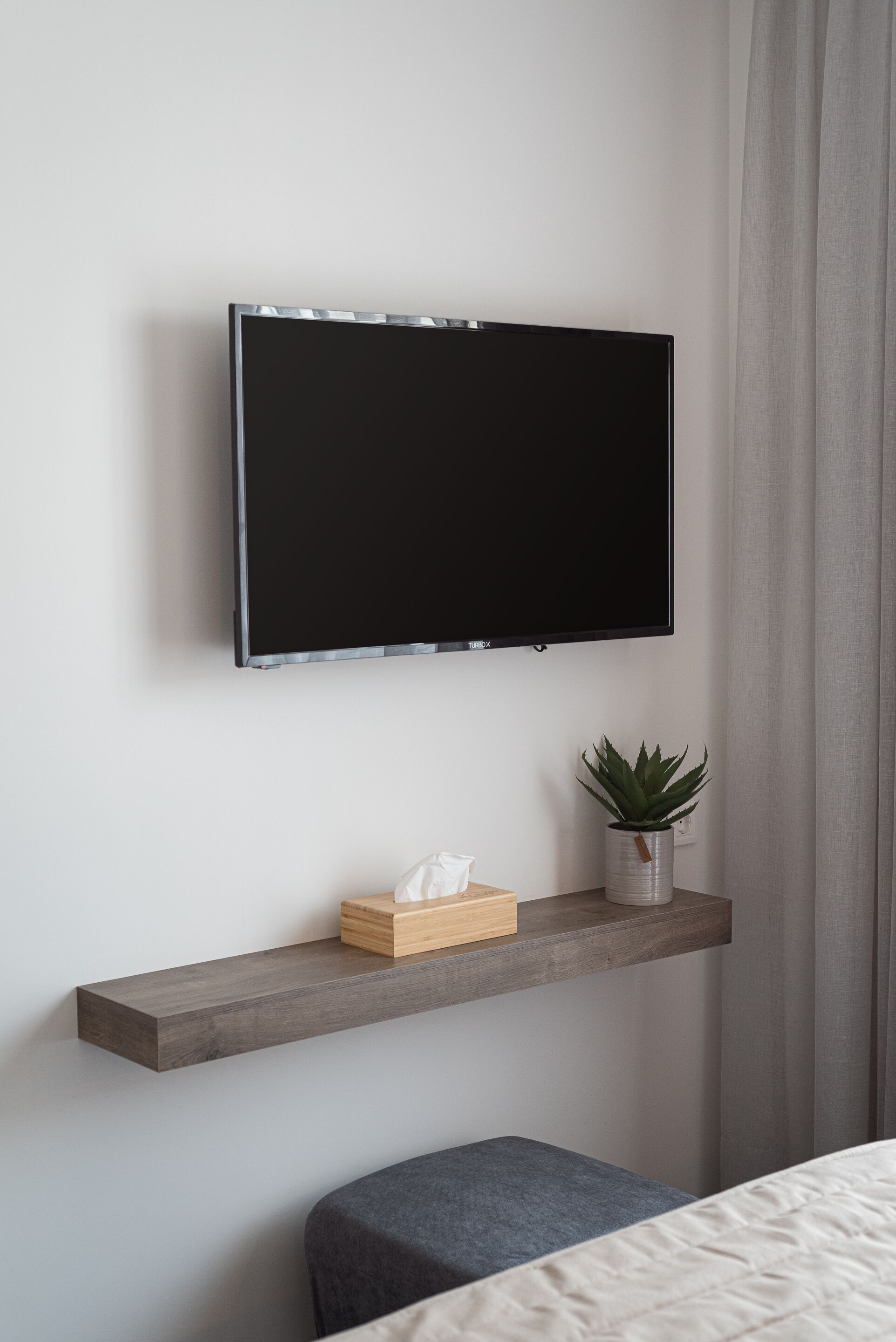 Ein modernes Schlafzimmer mit einem angebrachten Fernseher, einem Holzbodenregal mit Pflanze und Taschentüchern sowie einer grauen Bank.